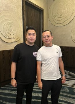 Two men standing side by side indoors, one wearing a black t-shirt with a small graphic and the other in a white t-shirt.