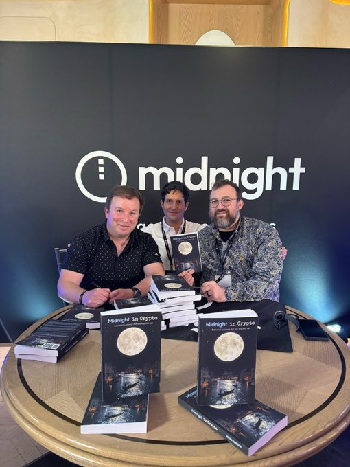 Three men pose at a round table in front of a "midnight" banner, with stacks of the book "Midnight in Orycto" displayed in front of them.