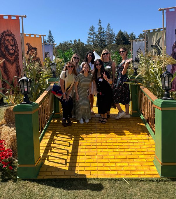 A group of eight women posing on a yellow brick road installation that resembles the Wizard of Oz, surrounded by corn stalks, hay bales, and Wizard of Oz character banners.