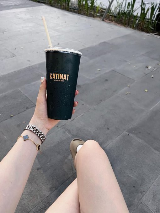 A hand wearing a clover charm bracelet holds a dark green "KATINAT" branded cup with a bamboo straw, while a leg in tan sandals rests on gray pavement nearby. The drink break creates a casual, relaxed moment that contrasts with the Game of Thrones-themed crypto musing shared in the caption.