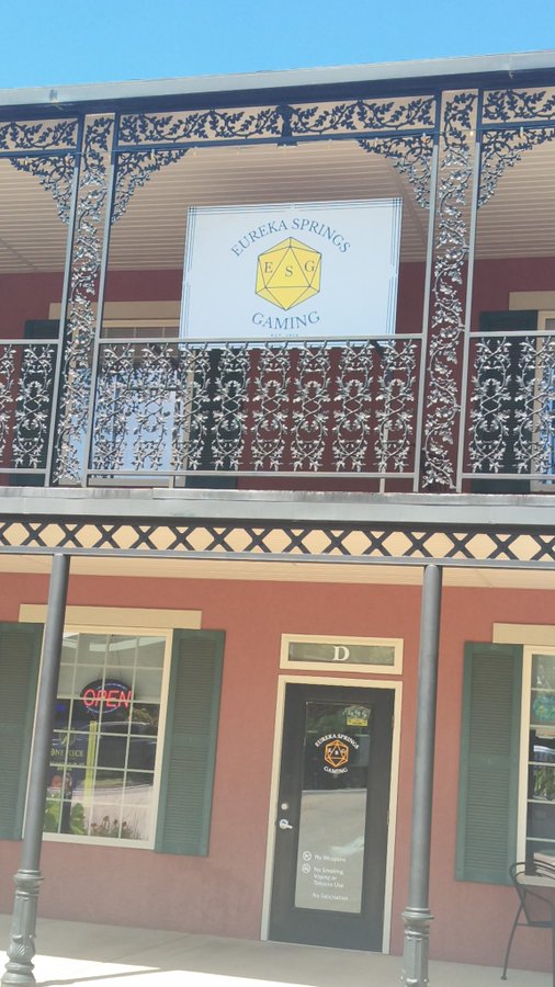 The storefront of 'Eureka Springs Gaming (ESG)' with an ornate iron balcony above, a red building facade, green shutters, and an 'OPEN' neon sign visible in the window.
