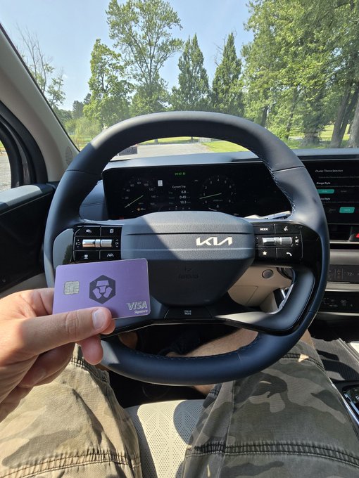A photo taken from inside a Kia vehicle showing the driver's hand holding a purple/pink Visa crypto card in front of the steering wheel.