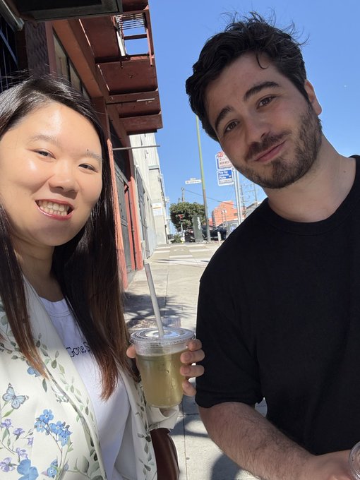 A smiling young Asian woman and a young man in a black t-shirt pose together outdoors on a sunny street, with the woman holding an iced coffee drink.