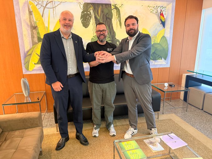 Three men stand together with their hands stacked in a gesture of partnership and unity in front of a tropical-themed mural, with one wearing a "Cardano" branded t-shirt. The image reflects the Cardano delegation's historic first stop at Petrobras headquarters in Brazil as part of their "Cardano on the Road" initiative aimed at connecting academia, industry, and government across Latin America.