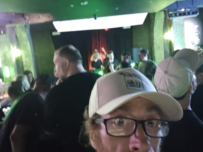 A selfie of a man wearing glasses and a white cap taken at a crowded nightclub or venue with green lighting.