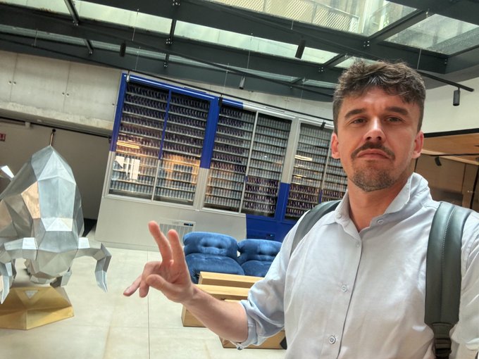 A man with a mustache wearing a light blue shirt and backpack gestures toward a metallic octopus sculpture in what appears to be a lobby or institutional building.
