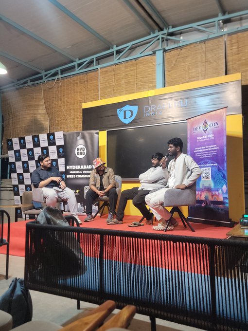 Four men sit on chairs on a stage during a panel discussion at 'DEVCON Hyderabad', with banners for 'Hyd Bro' and 'Dramified India' visible behind them.