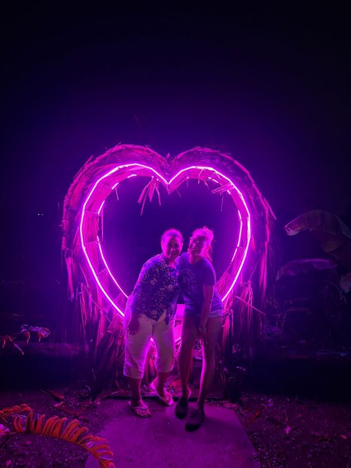 Two women pose together in front of a glowing pink neon heart-shaped installation in a dark, jungle-like setting.