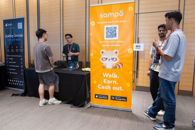 People gather at an indoor event booth for 'sampo - The easiest way to earn dollars' featuring a cartoon red panda mascot and the slogan 'Walk. Earn. Cash out.'