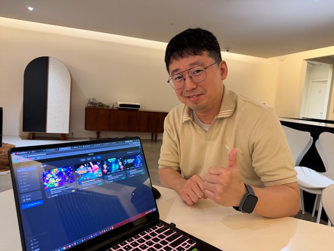 A man wearing glasses and a beige polo shirt smiles and gives a thumbs-up while sitting at a table with an open laptop displaying a colorful website.