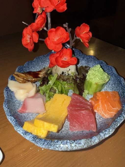 An artistically plated assortment of sashimi and sushi including tuna, salmon, and yellowtail, garnished with greens and decorative red flowers in a blue speckled ceramic bowl.