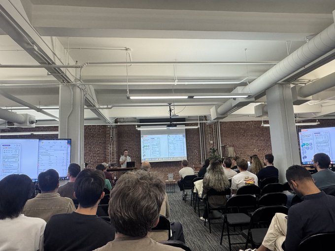 An audience sits in rows of chairs in an industrial-style room with exposed brick walls and pipes, watching a presentation being given at the front with two large screens displaying slides.