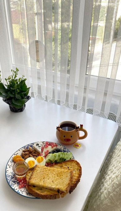 A cute breakfast spread featuring toast, a halved boiled egg, avocado slices, blueberries, almonds, and a character mug of coffee sits on a white table beside sheer curtains. The wholesome morning scene contrasts with the post's discussion about real-world asset infrastructure and @CNPYNetwork.