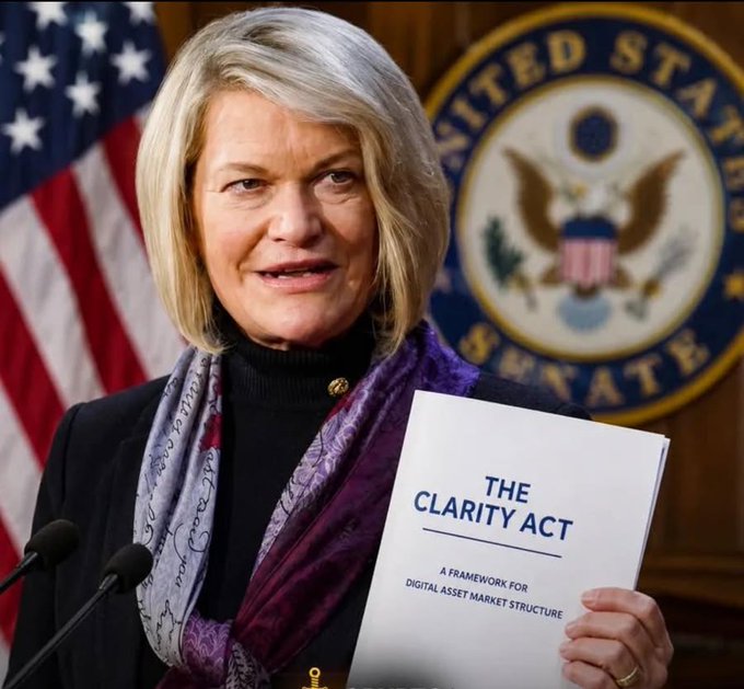 Senator Cynthia Lummis stands in front of a U.S. flag and the Senate seal, holding a document titled 'THE CLARITY ACT: A FRAMEWORK FOR DIGITAL ASSET MARKET STRUCTURE'.
