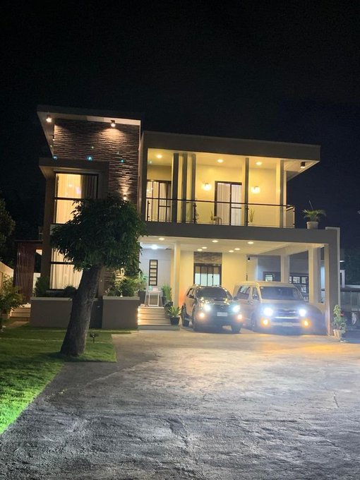 A modern two-story house illuminated at night with two cars (sedan and SUV) parked in the driveway, surrounded by a manicured lawn and small trees.