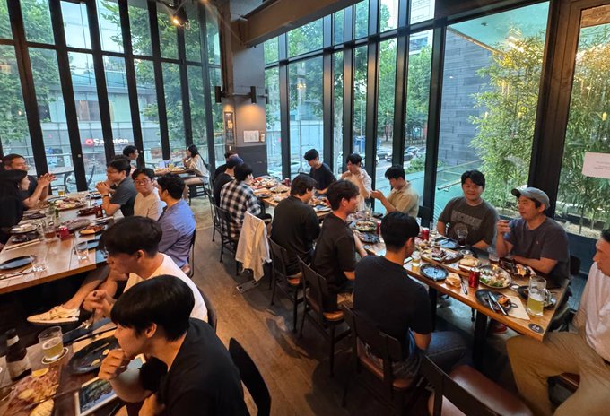 A lively dinner gathering of Solana ecosystem members, including core team members, developers, and founders, seated at long tables sharing food, drinks, and conversation in a bright, glass-walled venue. The scene captures the community spirit of reconnecting and aligning on shared goals, reflecting the post's emphasis on the real people driving the ecosystem forward together.