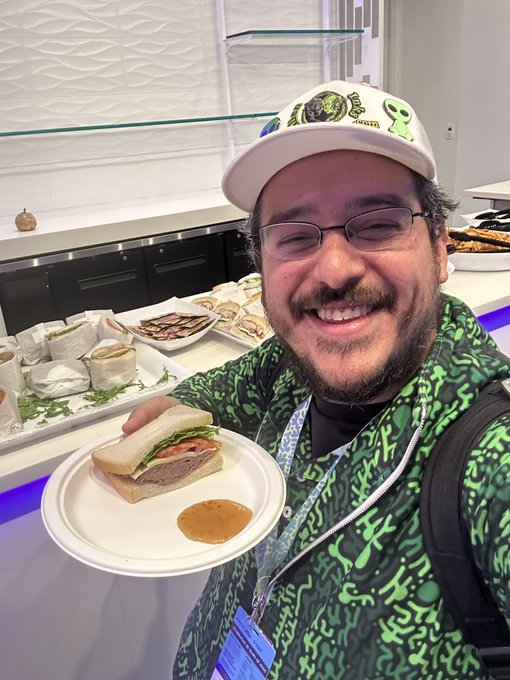 Bearded man wearing glasses and a white cap, smiling while holding a paper plate with a sandwich and dipping sauce at what appears to be an event or convention.