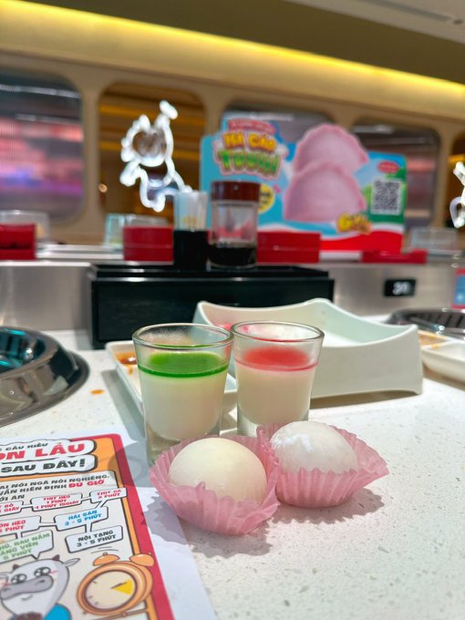 A photo of two small pastel-colored desserts (one green-topped, one pink-topped) in shot glasses next to two white mochi balls in pink wrappers, placed on a conveyor belt at a restaurant with a blurred 'Mochi Young' advertisement in the background.