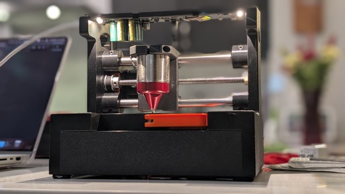 A compact CNC-style engraving machine, the SeedHammer, is shown in operation on a desk, with a red-lit tool head actively carving into a small orange workpiece. The setup sits beside a laptop and a vase of flowers, illustrating a hands-on testing session of the device.