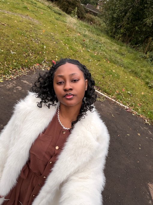 A woman with curly hair wearing a brown dress and fluffy white fur coat poses outdoors on a paved path for a selfie, exuding a confident and stylish vibe to greet followers with "Happy Tuesday."