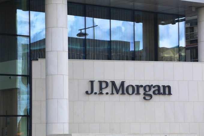 The exterior of a J.P. Morgan building displays the company's name in bold lettering on a light-colored stone facade, with large reflective windows above. This image relates to the post by visually representing J.P. Morgan, the financial institution highlighted for its rapidly growing treasury tokenization product that expanded to roughly 1.2 trillion.