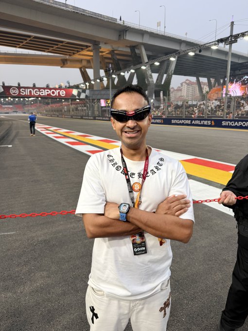 A smiling man wearing sunglasses and a "Chrome Hearts" t-shirt poses with crossed arms at the Singapore Grand Prix track, with "SINGAPORE AIRLINES" and "SINGAPORE" signage visible in the background.