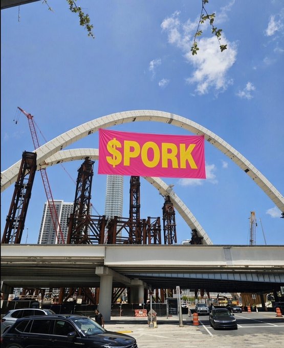 A photo of a large pink '$PORK' banner hanging beneath an arched overpass under construction, with cars driving below and cranes visible in the background.