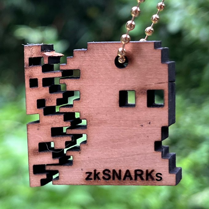 A wooden pendant shaped like a pixelated Minecraft-style creature hangs from a gold ball chain, engraved with the text 'zkSNARKs' against a green foliage background.