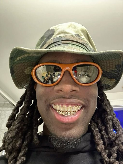 A close-up selfie of a person wearing a camo bucket hat, orange-tinted reflective sunglasses, and gold grills that spell out text on their teeth.