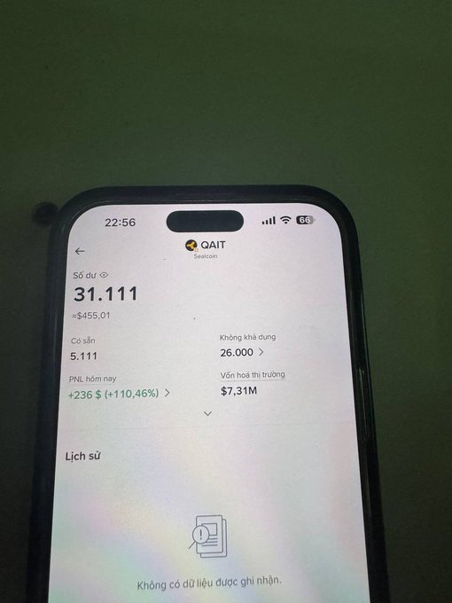 The image shows a cell phone screen displaying information about an item for sale, possibly a piece of jewelry or a collectible item. The text on the screen includes the name "QAIT Khá đậm khoảng 300$ nếu ae sell luôn lúc claim Mình đợt này hết điểm nên chỉ ngó thôi Ảnh coppy". The phone is placed on a table, and the background appears to be green.