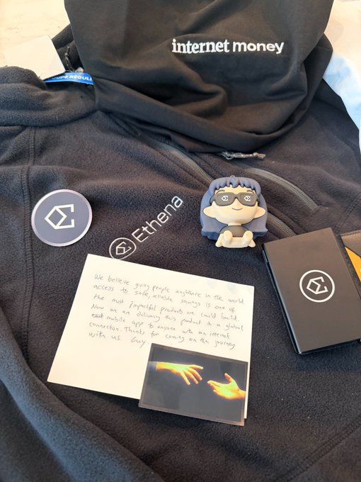 A collection of Ethena and "internet money" branded merchandise, including a black hoodie, stickers, a badge with a masked character, and a handwritten note alongside a "Creation of Adam" hands printout, visually reflects Ethena's mission of providing global, internet-accessible savings as quoted in the post.