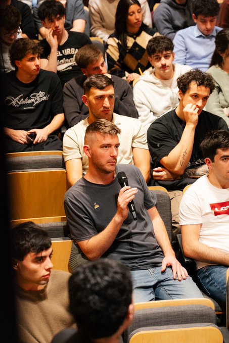 A young man in a gray t-shirt holding a microphone while seated in an auditorium filled with attentive audience members listening to a presentation.