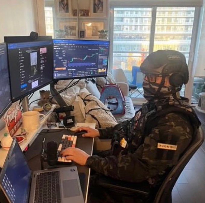 A person in tactical gear, likely trading cryptocurrency, sits at a desk with multiple monitors displaying financial charts and data. The image humorously depicts the intensity and preparedness some traders adopt, even with a small portfolio.