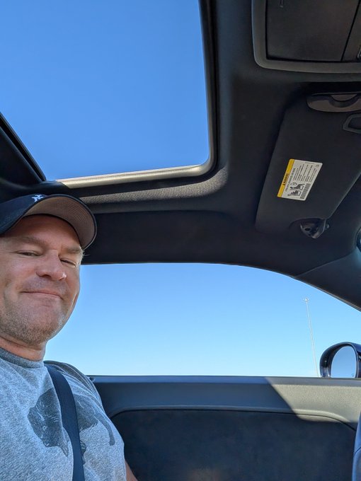 A selfie of a man wearing a dark baseball cap and grey t-shirt, smiling while sitting in a car with an open sunroof under a clear blue sky.