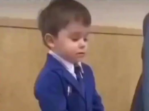 A young boy with brown hair wearing a blue blazer and tie, appearing to have his eyes closed or be looking down.