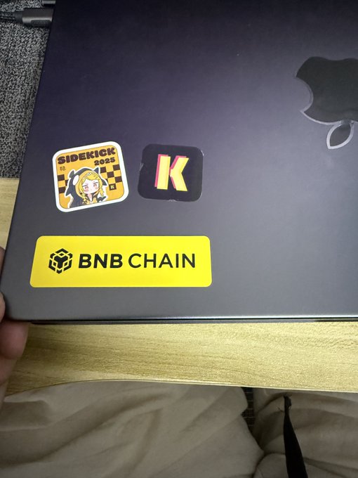 A dark gray laptop with Apple logo, displaying stickers including 'BNB CHAIN', a 'K' sticker, and a 'SIDEKICK 0025' anime character sticker.
