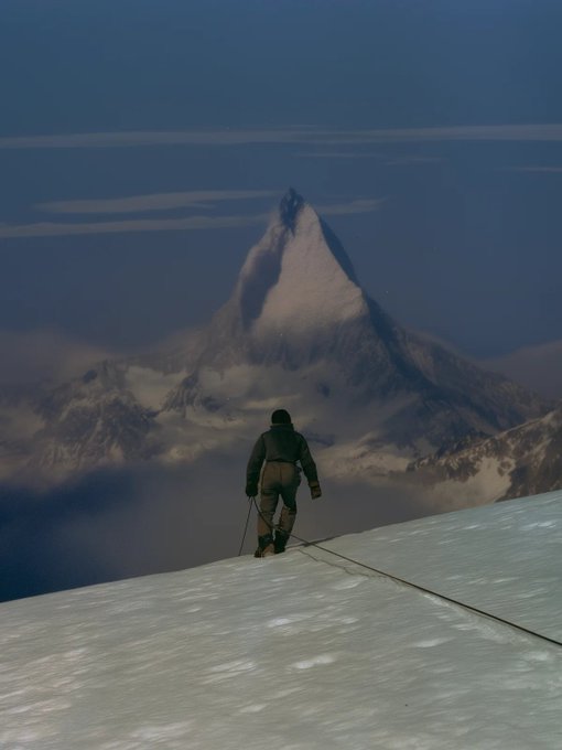 A lone climber stands on a snowy ridge with ice axe in hand, gazing toward the iconic Matterhorn peak piercing through clouds at dusk. The image metaphorically reflects the post's theme of an arduous journey forward—mirroring the slow, seven-year evolution of "Unfinished Past" on SuperRare, where each collector's choice to remember or let go shapes the path ahead.