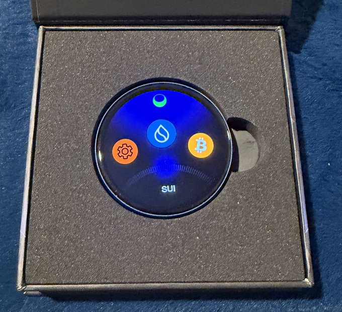 A circular SUI cryptocurrency hardware wallet rests inside its black packaging box, displaying icons for SUI, Bitcoin, Sui, and settings on its lit-up screen.