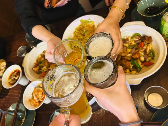 Friends clink mugs of cold beer over a spread of Chinese dishes, including stir-fried vegetables and meat with peppers, celebrating a lively evening out where the night has only just begun.