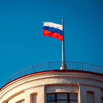 The Russian tricolor flag flies atop a domed building against a clear blue sky, visually representing the country referenced in the news of Sberbank's $46.4 billion forecast for Russia's regulated crypto trading volume.