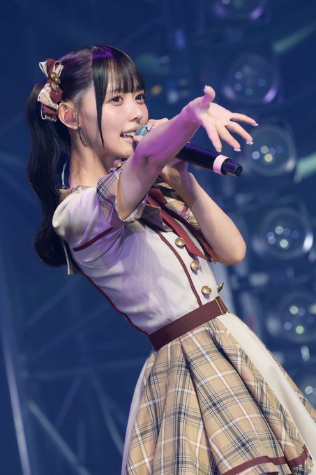 A young Japanese idol with pigtails performs on stage, singing into a microphone while wearing a white and plaid school-style dress.