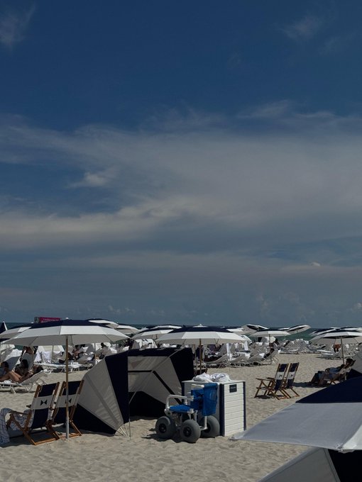 A crowded beach scene with numerous white umbrellas, beach chairs, a blue ATV, and people under a partly cloudy blue sky.