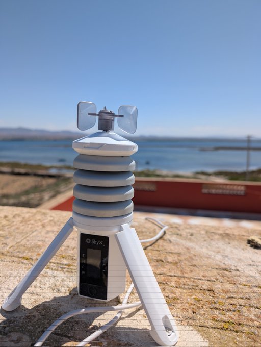 The image shows an electronic device with a white base that has been placed outdoors on a rooftop or balcony. The device appears to be a weather station or sensor, possibly used for measuring wind speed and direction. It is connected to a power source via a cord, indicating that it is operational and ready for use.