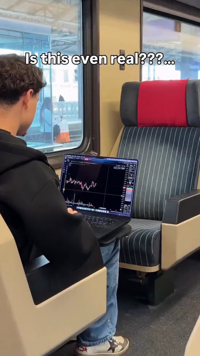 A young man sits on a train trading stocks or crypto on his laptop, with overlaid text reading 'Is this even real???...'