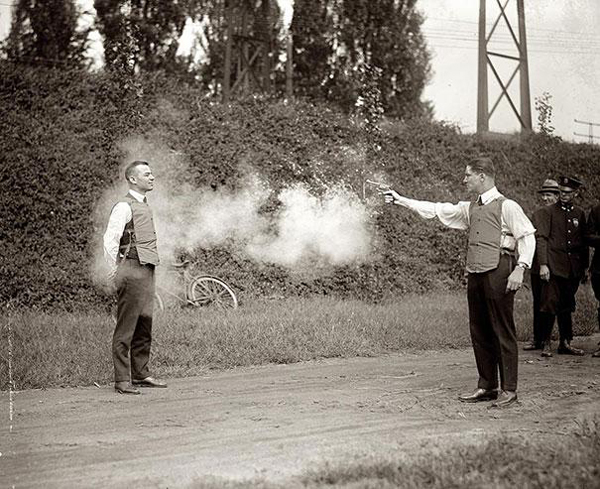 A black and white historical photograph showing a man being shot with a pistol by another man while officers look on outdoors.