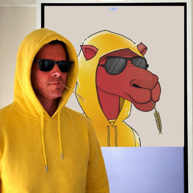A man in a yellow hoodie and sunglasses holding a wheat stalk in his mouth, standing next to a framed cartoon artwork of a camel in a matching yellow hoodie and sunglasses.