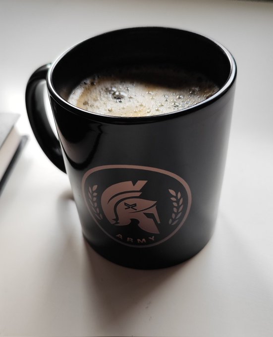 A black mug filled with frothy coffee sits on a white surface, featuring a copper-colored Spartan helmet emblem with the word "ARMY" beneath it. The mug visually pairs with the "Gm fam!" post by representing the coffee in hand, complementing the reference to holding $XRP in the other.