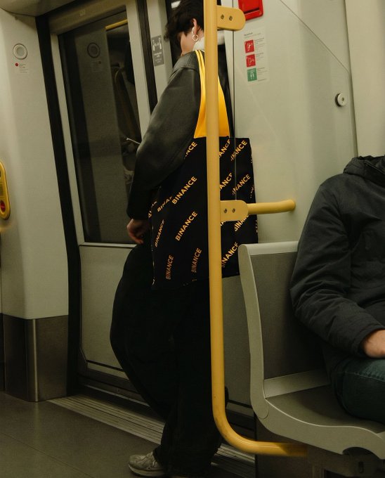 The image shows a person standing in the doorway of a subway car holding a yellow handbag with the Binance logo. The accompanying post says "volviendo..." which is Spanish for "coming back," possibly indicating that the person has finished their errands or outing and is on their way back home.
