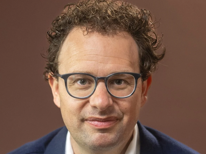 A close-up portrait of a curly-haired man with dark-rimmed glasses, wearing a navy blazer, smiling slightly against a brown background—likely Dario Amodei, CEO of Anthropic, the AI company whose year-end valuation prospects are being speculated about.