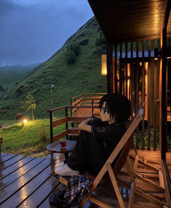 An anime-style illustrated character with dreadlocks sitting in a wooden chair on a porch, overlooking a grassy mountainside with a cow and misty hills in the background at dusk.